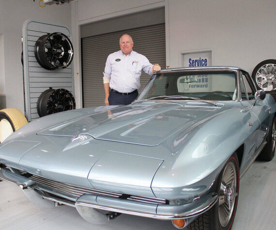 <p>Alan Gamblin, who took over his father’s dealership in 1985, stands in his car shop next to his personal 1964 Corvette in 2019 while celebrating being 50 years in business on the Plateau. Photo by Ray Miller-Still</p>